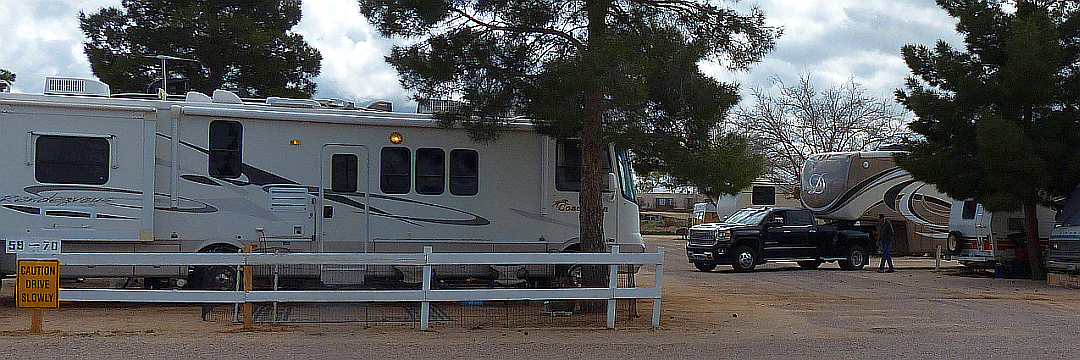 A motorhome parked under tall pines beside the campground's white rail fence